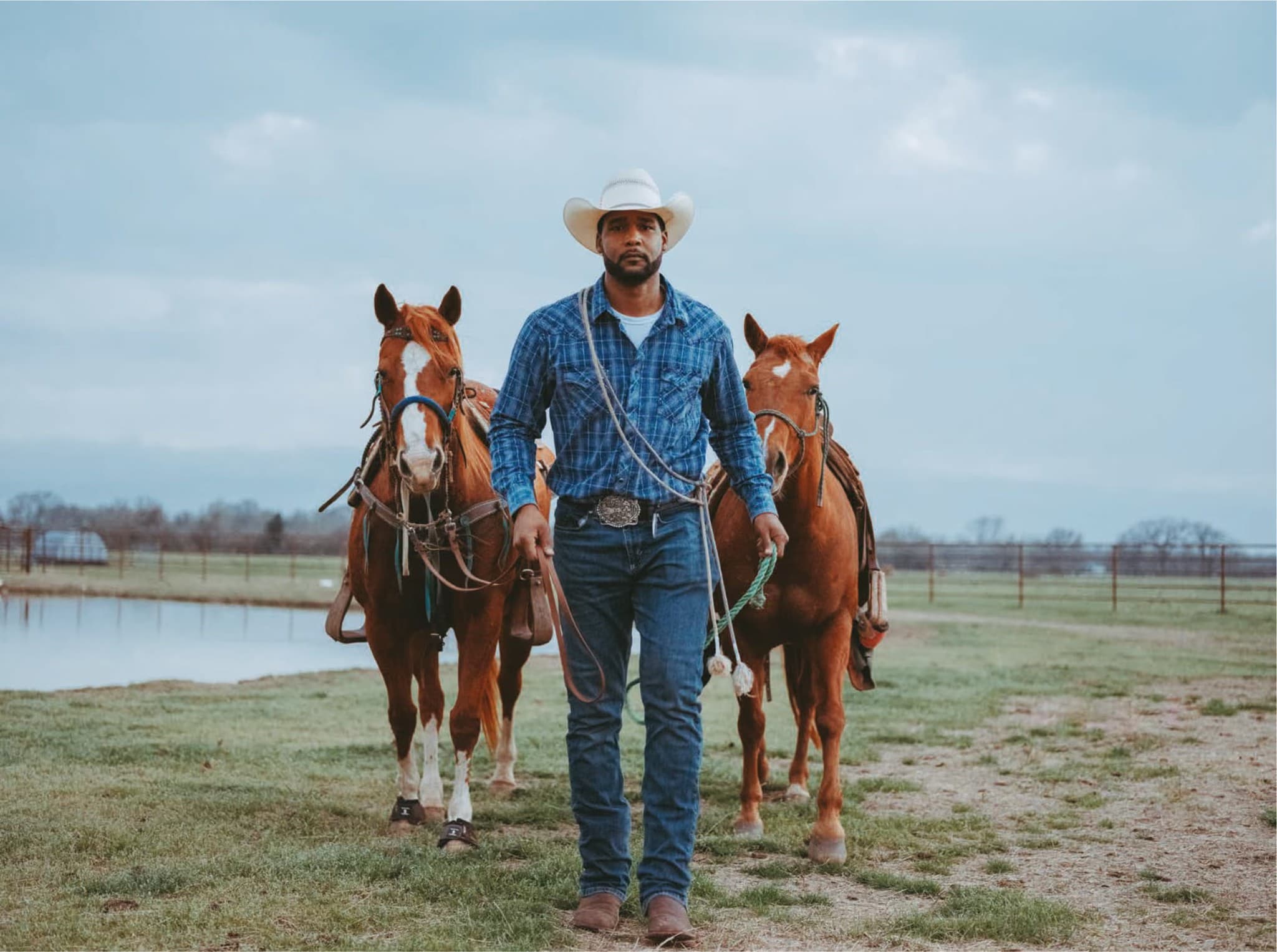 Black cowboy in Western attire with two horses in a grassy field